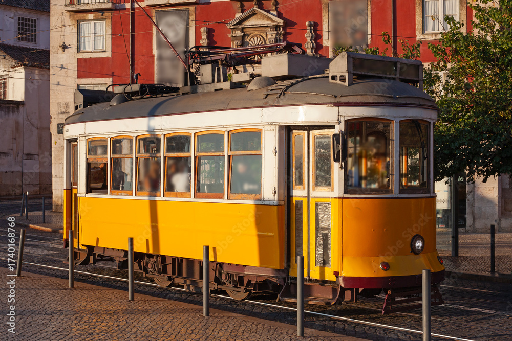 Naklejka premium Lisbon, Portugal - July 27, 2025: A classic fampus yellow tram passes by vibrant red historic buildings in Lisbon early sunrise morning