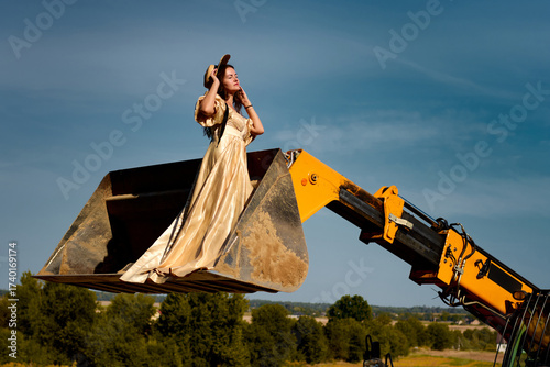 Tableau sur toile A beautiful woman in an opulent golden dress sits regally in a backhoe bucket against a clear blue sky and lush green landscape, creating a surreal and striking juxtaposition