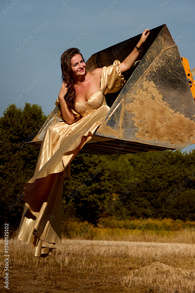 Fototapeta premium Elegant woman in golden dress confidently poses inside an excavator bucket in field, creating a striking contrast between glamour and rugged machinery, radiating beauty and empowerment in nature.