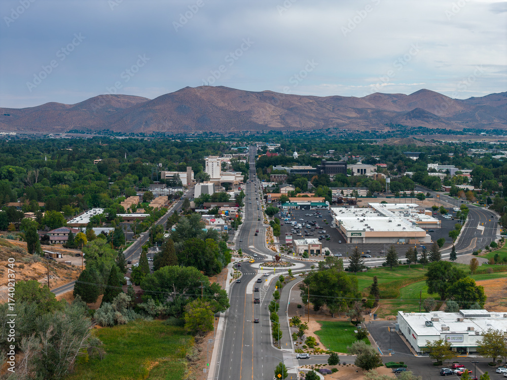 Fototapeta premium Aerial view of Carson City, Nevada, showing a central road, commercial buildings, green spaces, trees, arid hills, and distant mountains.