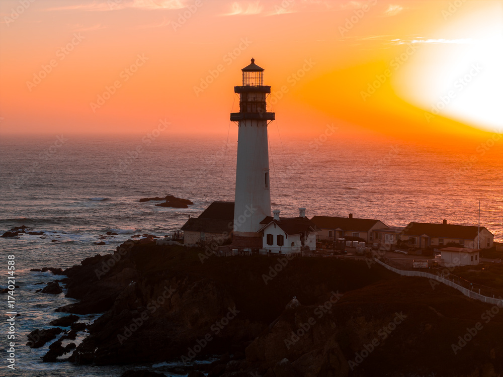Naklejka premium Aerial view of Pigeon Point Lighthouse on a coastal cliff in California, surrounded by ocean waves, rugged rocks, and warm sunset hues.
