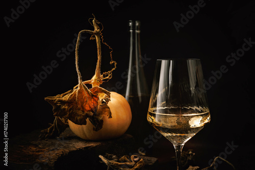 Glass of white wine with pumpkin bottle and dried leaves on rustic wooden surface in dark setting