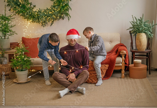 The children tie up their father, dressed up as Santa Claus, with a garland of lights. Funny, humorous boys are joking about Dad. On the eve of Christmas and New Year