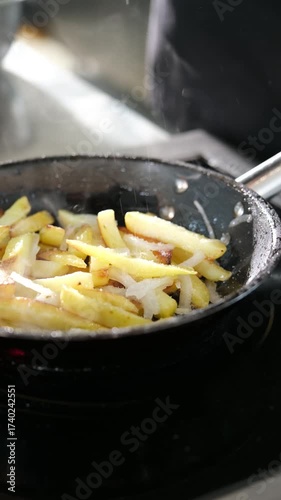Professional chef salting steaming hot french fries and sliced onions cooking in a black frying pan on an electric stove, creating a delicious and unhealthy fast food side dish. Vertical video