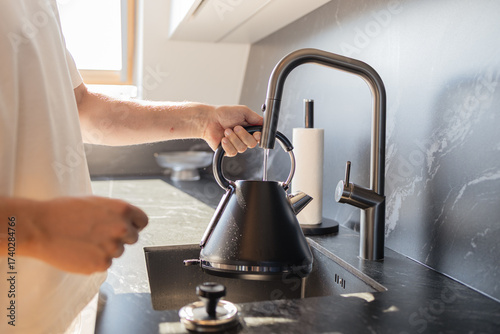 Close-up of man pouring water into black kettle from stylish faucet in modern kitchen. Concept of daily routine, home lifestyle, and cooking preparation. Symbol of household comfort, minimalistic