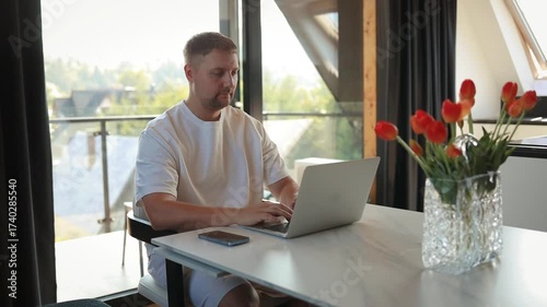 Casual man sitting at desk with laptop in bright home interior, working remotely or studying online. Concept of freelancing, digital lifestyle, remote job, technology, communication and productivity