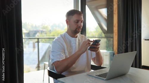 Young man sitting at desk with computer and phone, paying bills or shopping online. Cashless economy, mobile banking, household budget, financial technology and remote work.