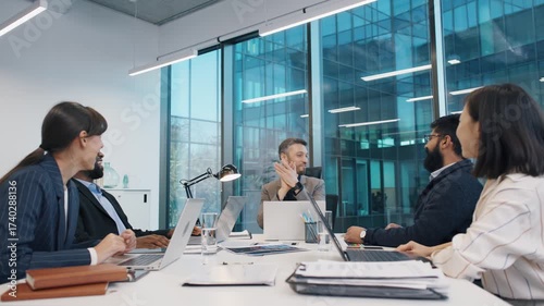 Business team laughing together during relaxed strategy session in modern glass office, led by man in grey blazer. Diverse professionals seated around laptops and charts, exchanging ideas.