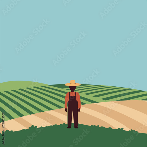 A person in overalls and a hat stands on a grassy hill overlooking striped agricultural fields under a blue sky
