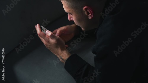 A man types on a mobile phone while lying on a couch in a room.
A stylish man holds a black touchscreen phone while sitting on a sofa in a room.
