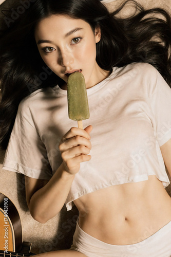 a person lying on the ground, wearing a white crop top and underwear, holding a green popsicle. They are surrounded by a guitar, an open book, and another book on the ground. The setti
