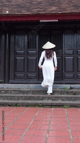 
4k Vertical video.Woman in traditional Vietnamese áo dài and conical hat walking in front of ancient wooden door, symbol of Vietnam culture and travel lifestyle.