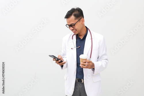 Asian adult male doctor smiling wearing doctor's coat and hanging stethoscope on neck isolated on white background holding paper cup of coffee while using mobile phone.