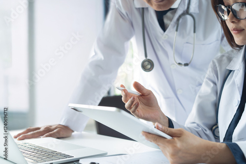 Two asian doctors discuss diagnosis, analyzing data on digital tablet in hospital office. Male and female medical professionals have a patient case conference, using tablet and laptop, close up