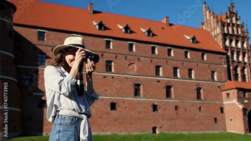 Happy stylish female tourist in a hat smiling and taking photographs of a historic red brick castle on a sunny summer day.