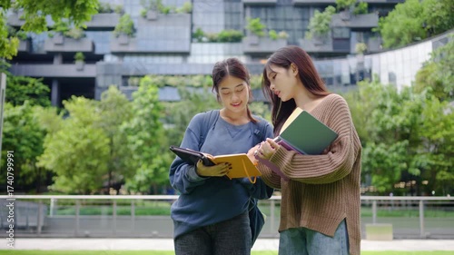 Two young Asian female students enjoy a lively study session outside their university. They exchange ideas, smile, and laugh, showcasing the joy of learning together in a beautiful green setting.