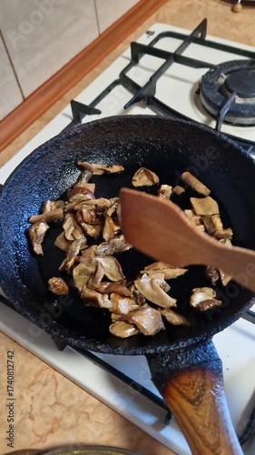Close-up shot a hand stirring chopped brown mushrooms in a dark cast iron frying pan on a white gas stove, preparing a meal in a home kitchen environment, showcasing culinary process.