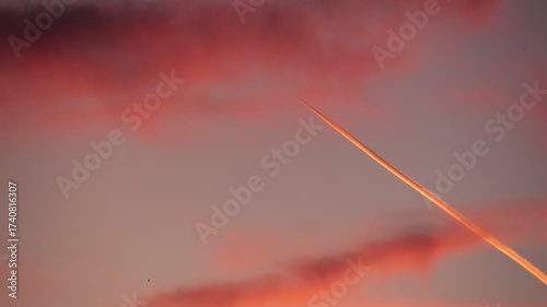 A vibrant sunset sky with pink and orange clouds. A jet contrail cuts through the sky, creating a striking contrast against the colorful backdrop.