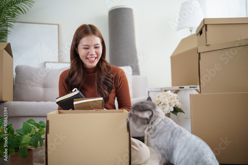 Young woman unpacking boxes while moving into a new home, smiling and playing with her cat in a cozy living room.