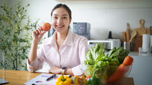 Smiling female nutritionist holding a fresh apple while sitting at desk with vegetables and fruits. Healthy food and diet planning concep