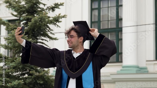 Modern graduate boy with cap on his head and graduation gown poses and takes selfies on his phone to send to his parents and friends. Pictures from the graduation ceremony for graduating students