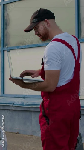 Technician in red overalls using diagnostic computer to troubleshoot engine issues, standing beside open hood of vehicle in a well-lit automotive workshop environment