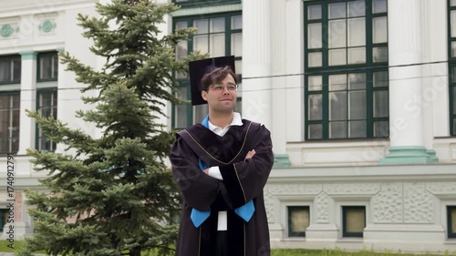 Handsome dark-haired graduate in a cap and black robe, confident and proud, nods demonstratively as a sign that he has succeeded. Boy with glasses at the university graduation ceremony