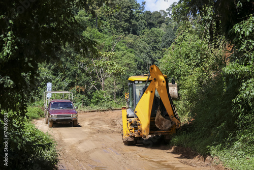 Yellow backhoe loader works on dirt road construction in rural jungle. red pickup truck drives on unpaved path showing progress and development in remote green forest