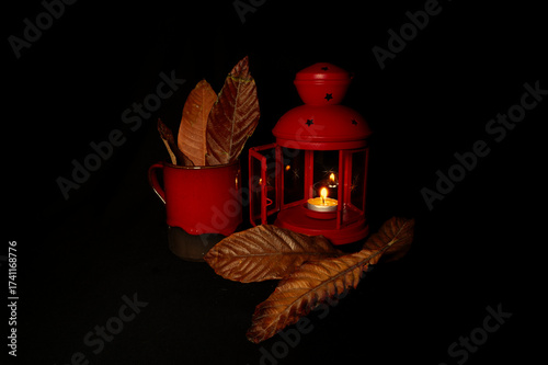 utumn still life with red cup, lantern, candle and dry leaves on dark background. Warm seasonal mood evoking fall evenings and cozy atmosphere, also suitable for Halloween themes.