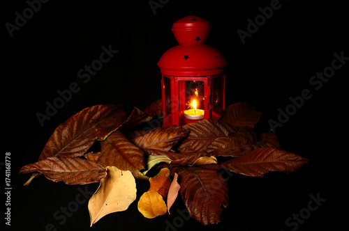 Red lantern with a burning candle surrounded by dry autumn leaves on dark background. Warm seasonal still life evoking fall evenings, cozy atmosphere and mysterious Halloween mood