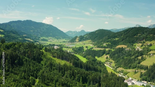 An epic aerial view of the vast mountain landscape of the Austrian Alps. A drone flies over a wide green valley with forests, hills, and rural villages, a beautiful summer travel destination.