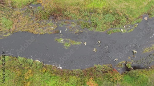 Aerial drone view of a winding river flowing through a green countryside landscape. Top-down perspective showing curved waterway with autumn foliage and grass banks