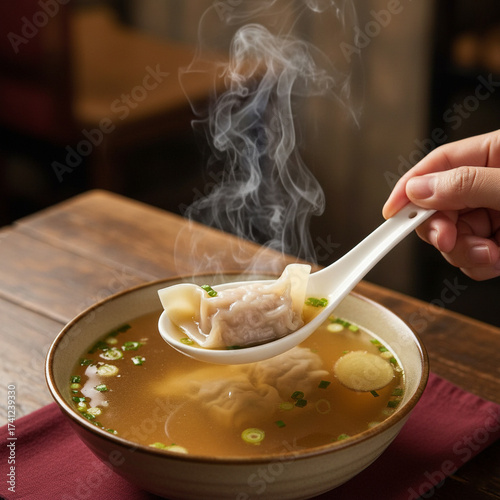 hand holding a porcelain spoon lifted slightly above a bowl, containing a perfectly folded wonton submerged in clear, steaming broth