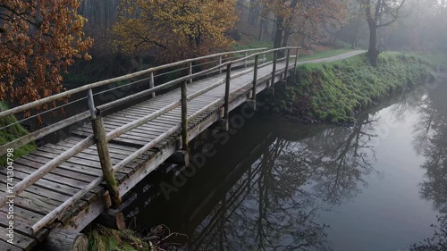 Old wooden bridge crossing foggy autumn river surrounded by hazel trees and misty countryside landscape