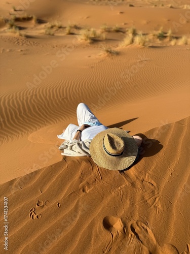 A woman in a hat lies on a sand dune in the Sahara Desert in Merzouga. Perfect for use in publications or as a design element that symbolizes freedom, emptiness, and nature