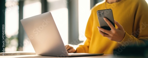The laptop and smartphone on a sunlit desk with a person using mobile