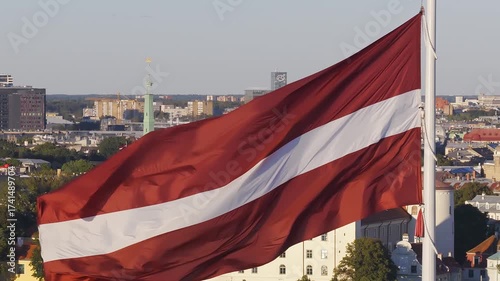 Aerial view of Riga, Latvia, featuring the waving national flag, historic old town with St. Peter's Church spire, modern buildings, and greenery.