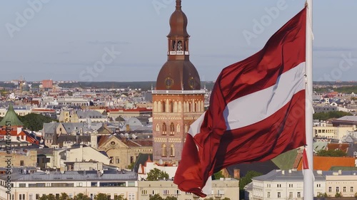 Aerial view of Riga, Latvia, featuring the national flag waving in the wind, Riga Cathedral's spire, and historic old town architecture under clear skies.