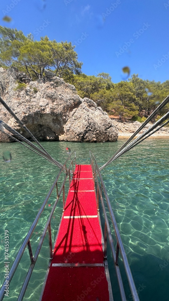 Fototapeta premium Red-carpeted boat ramp leading down into the crystal clear, shallow turquoise water toward a rocky, tree-covered cove in Adrasan