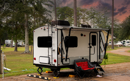 A small white camper sits in a tranquil RV park surrounded by tall trees. The setting sun casts a warm glow over the scene, 