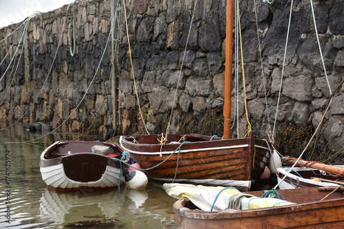 The image shows small wooden boats tied with ropes along a stone harbor wall. The boats float in calm, shallow water, giving a rustic and peaceful coastal atmosphere.