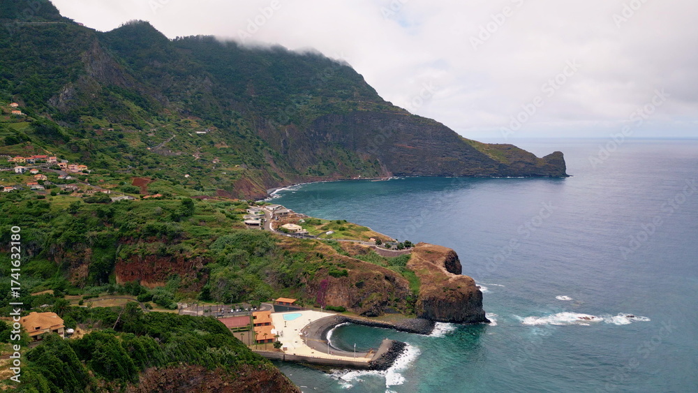 Fototapeta premium Ocean water washing green mountain slopes with cozy small village drone view. 