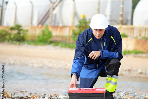 Environmental engineer or ecological scientist industrial worker in safety uniform working for inspect water quality at industry construction site near oil refinery plant by the sea.