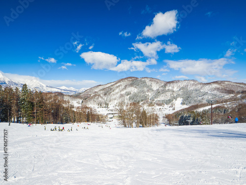 A wide ski slope with many people skiing and snowboarding on a sunny winter day (Madarao, Nagano, Japan)