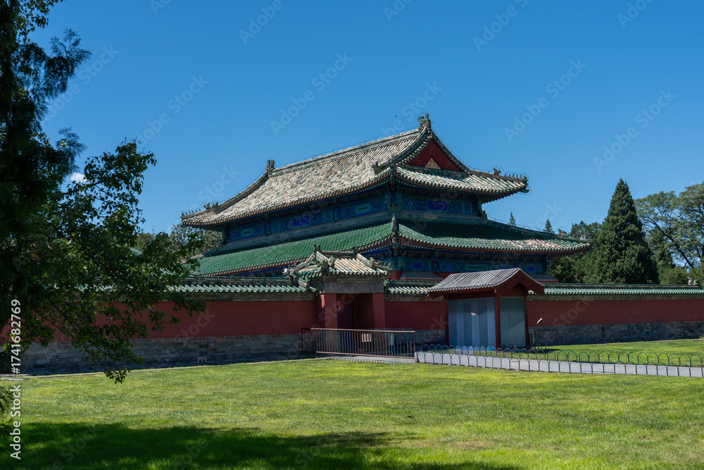 Obraz premium A majestic traditional Chinese temple or hall with an intricate green tiled roof and a perimeter red wall. The historic structure is set against a blue sky and green lawn, conveying culture