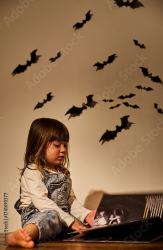 Charming child or serious girl in overalls reads a Halloween book under warm lighting, with bat wall decorations creating a cozy, spooky ambiance. Autumn, storytelling, and spooky festive concept
