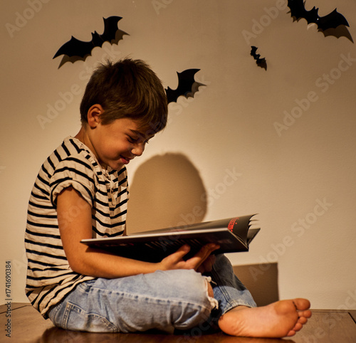 Enthusiastic child or smiling boy reading a book in cozy Halloween setting with bat decorations on wall. Warm lighting creates festive, spooky autumn atmosphere. Perfect for seasonal education themes