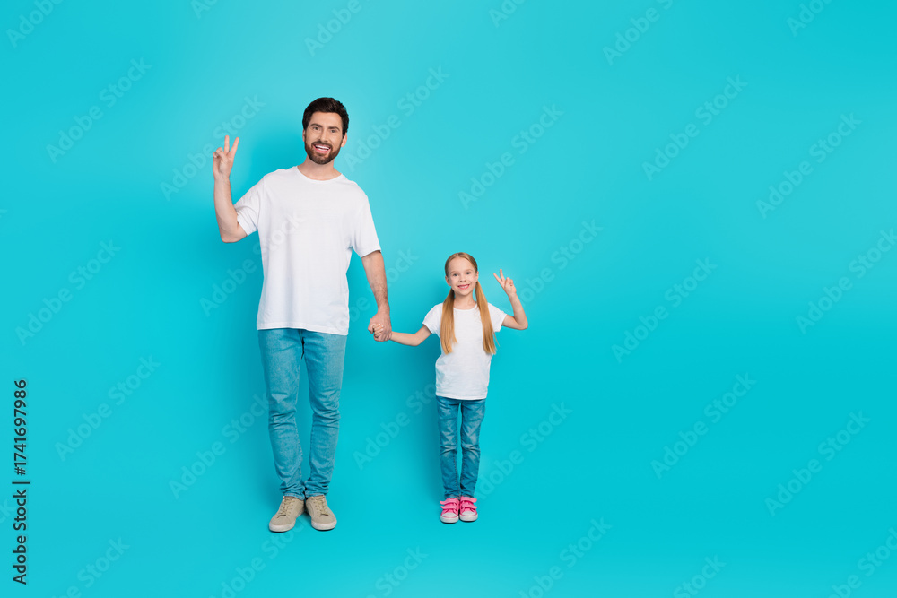 Fototapeta premium Dad and daughter wearing white t-shirts showing peace gesture against a blue studio backdrop, displaying joy
