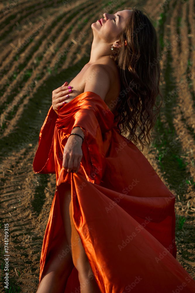 Fototapeta premium A beautiful young woman dressed in an orange dress, she stands in a vast plowed field, with her face turned upwards, enjoying the warmth of the sun and the fresh air, feeling the serenity of the