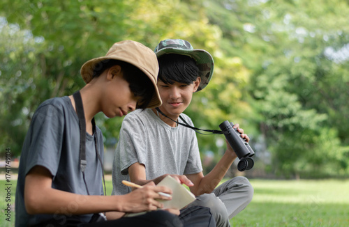 Two Asian boys exploring nature, one using binoculars and the other writing notes. Outdoor learning, curiosity, and childhood adventure in a green, forest-like environment.